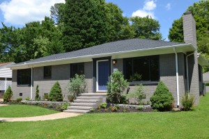Gray painted brick home with blue front door black trim windows and frame around door steps to front door and bushes and flowers in front yard