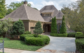 Brown shingle brick and stone two story home with high sloped brown shingle roof brown two car garage door blue shutters manicured trees and bushes