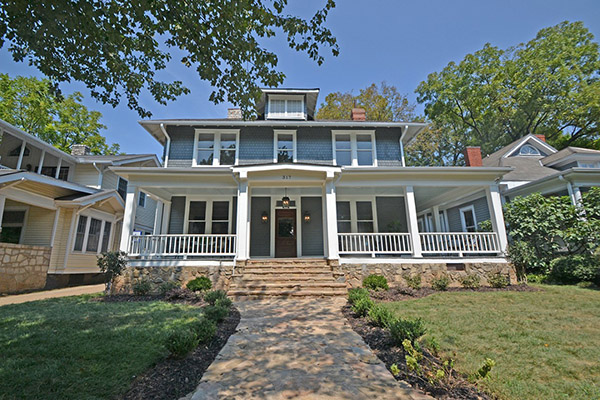 Three story home with gray siding and gray shingles brown front door front porch with white columns and white railing stone detail along bottom